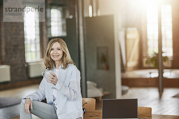 Lächelnde Frau mit Kaffeetasse sitzt zu Hause am Tisch