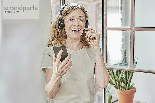 Glückliche Frau mit Headset und Smartphone vor dem Fenster zu Hause