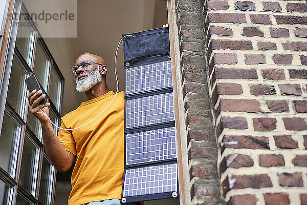 Mann mit Smartphone und Solarpanel steht zu Hause am Fenster