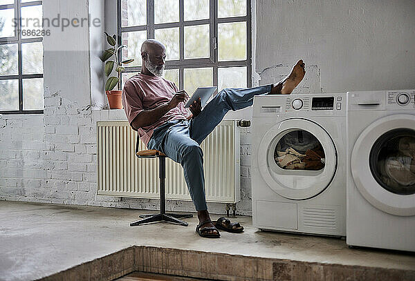 Reifer Mann benutzt Tablet-PC mit Fuß auf Waschmaschine und sitzt zu Hause auf einem Stuhl