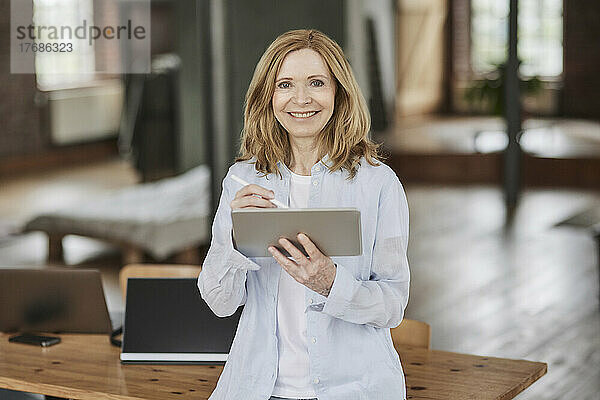 Lächelnde Geschäftsfrau mit Tablet-PC  die sich zu Hause auf den Tisch lehnt