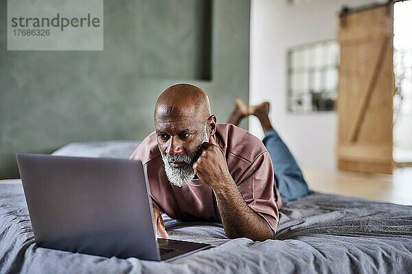Mature man with white beard using laptop lying on bed at home