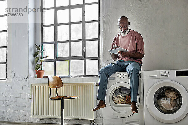 Mann mit Glatze benutzt Tablet-PC und sitzt zu Hause an der Waschmaschine