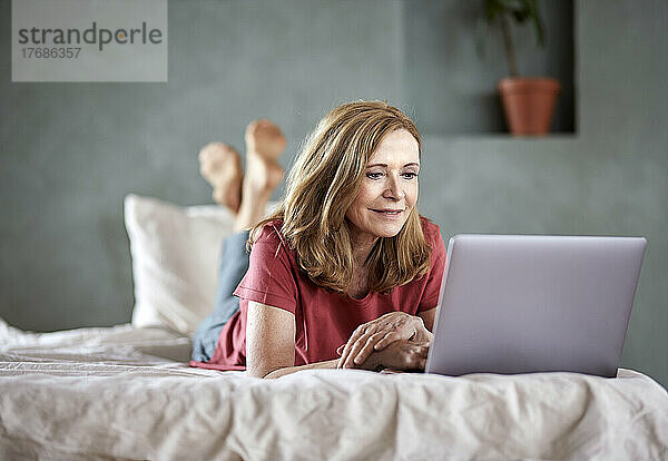 Lächelnde Frau mit Laptop  die zu Hause im Bett liegt