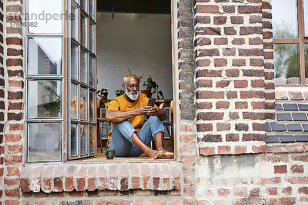 Reifer Mann surft im Internet über sein Mobiltelefon und sitzt am Fenster
