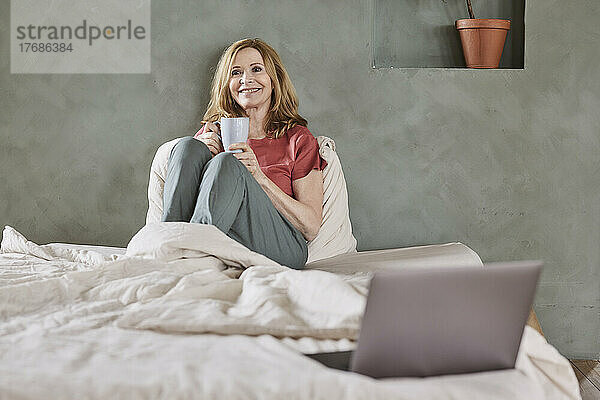 Lächelnde Frau mit Kaffeetasse sitzt zu Hause im Bett