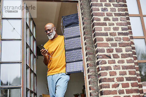 Mann mit Glatze benutzt Smartphone und steht mit Solarpanel am Fenster zu Hause