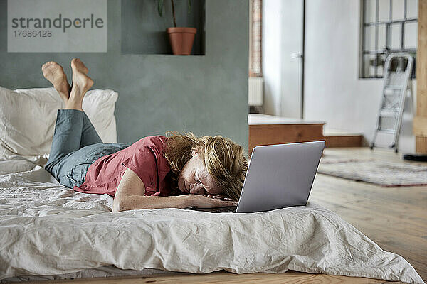 Tired woman lying by laptop on bed at home