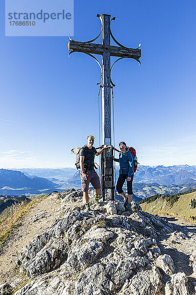 Deutschland  Bayern  Zwei Wanderer posieren vor dem Gipfelkreuz am Geigelstein