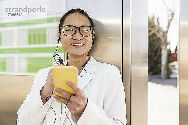 Lächelnde Geschäftsfrau hält Smartphone in der Hand und hört Musik über In-Ear-Kopfhörer