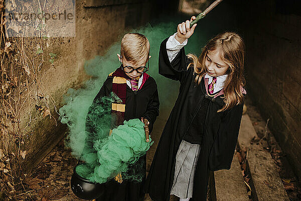 Mädchen mit Bruder hält Kessel und Zauberstab in der Hand und steht inmitten von grünem Rauch im Tunnel