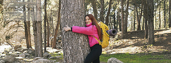 Glückliche junge Frau mit Rucksack umarmt Baumstamm im Wald