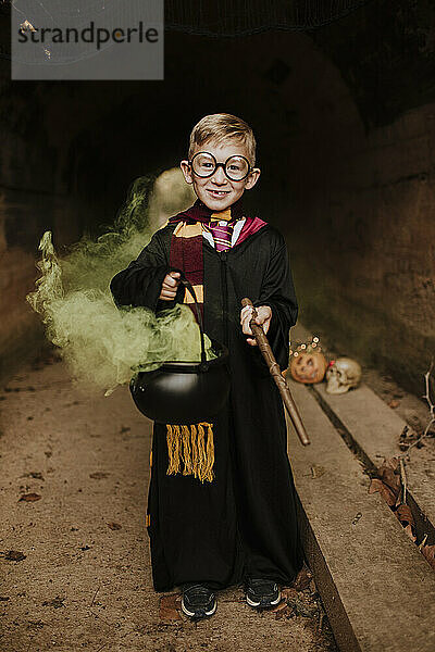 Lächelnder Junge hält einen magischen Kessel mit grünem Rauch und trägt ein Kostüm im Tunnel