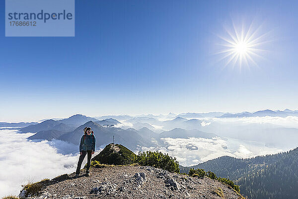Deutschland  Bayern  Sonne scheint über einer Wanderin  die auf einem Berggipfel steht  mit dichtem Nebel und dem Gipfel des Herzogstands im Hintergrund