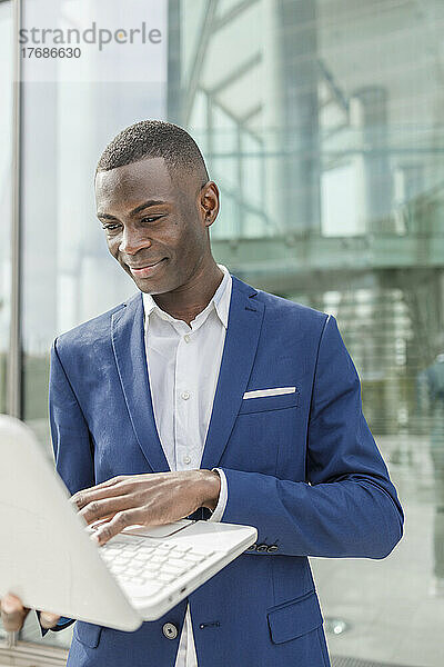 Lächelnder Geschäftsmann mit Laptop vor modernem Bürogebäude
