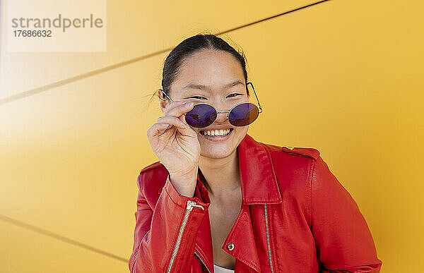Glückliche junge Frau mit Sonnenbrille steht vor gelber Wand