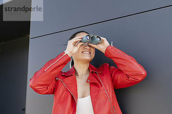 Smiling young woman looking through binoculars in front of gray wall