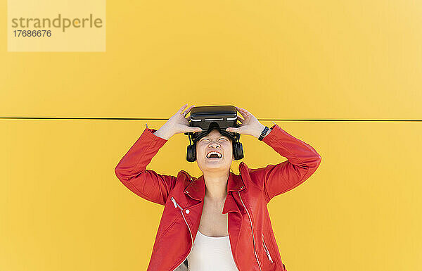 Glückliche Frau hält Virtual-Reality-Simulator vor gelber Wand