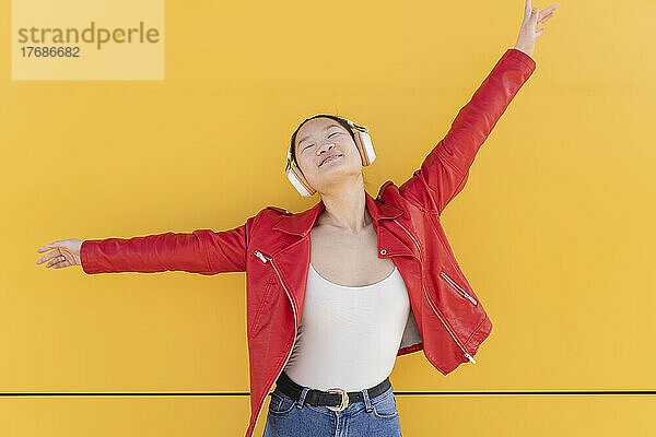 Young woman listening music through wireless headphones in front of yellow wall