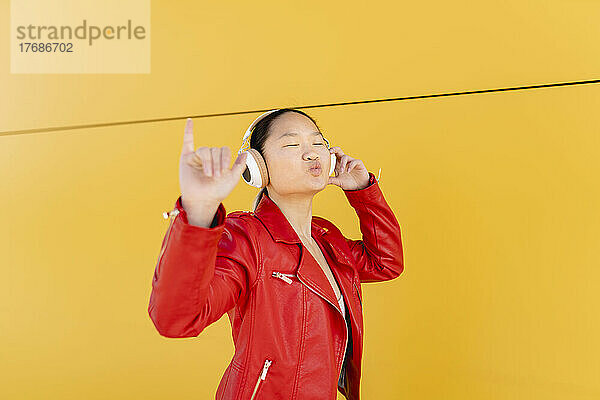 Junge Frau hört Musik über drahtlose Kopfhörer und zeigt Shaka-Schild vor gelber Wand