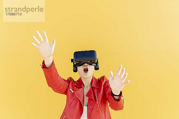 Junge Frau mit Virtual-Reality-Simulator gestikuliert vor gelbem Hintergrund