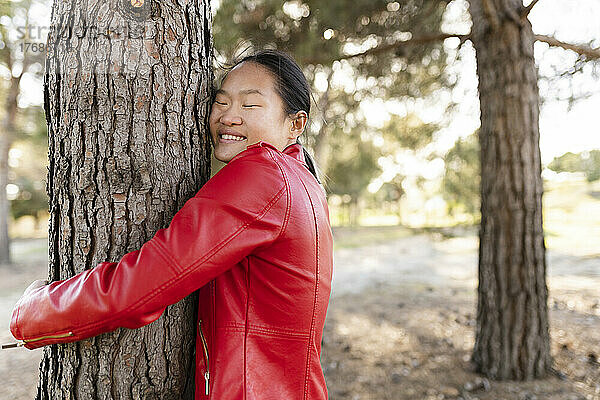 Glückliche junge Frau in roter Jacke umarmt Baum im Wald