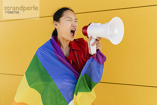 In Regenbogenfahne gehüllte Frau schreit durch Megafon an gelber Wand