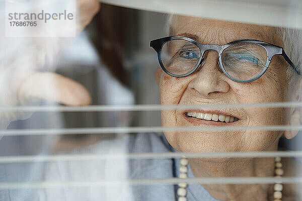 Happy senior woman looking through window blinds