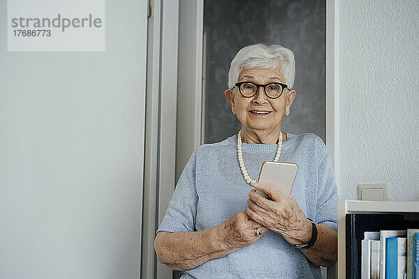 Lächelnde ältere Frau mit Mobiltelefon steht zu Hause