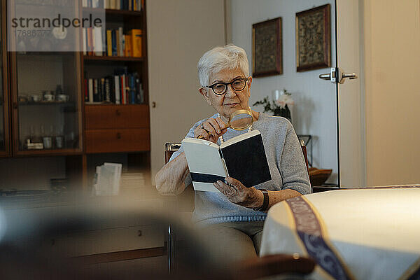 Ältere Frau liest zu Hause Buch mit Lupe