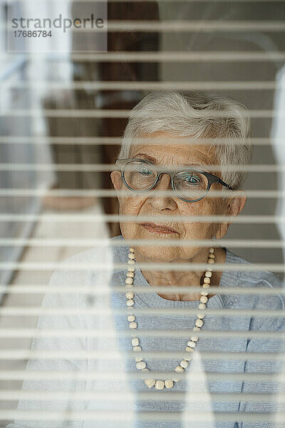 Ältere Frau im Ruhestand durch Fensterjalousien gesehen