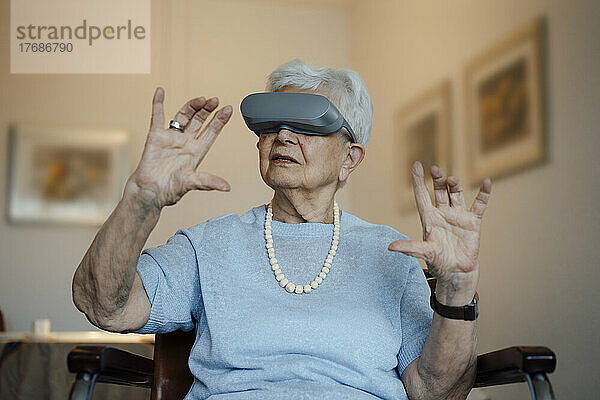 Ältere Frau mit Virtual-Reality-Simulator sitzt zu Hause auf einem Stuhl und gestikuliert