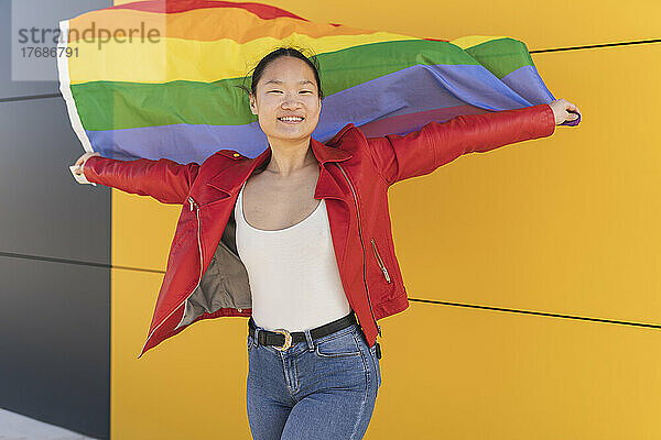 Lächelnde Frau mit Regenbogenfahne vor gelber Wand