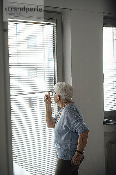 Ältere Frau blickt zu Hause durch die Jalousien nach draußen