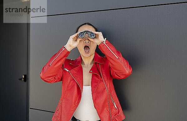 Schockierte Frau mit offenem Mund blickt durch ein Fernglas vor einer grauen Wand