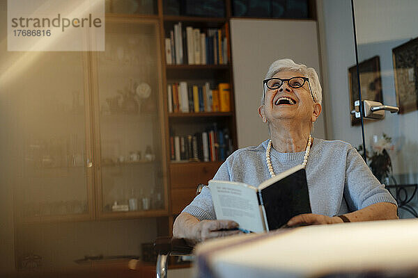 Happy senior woman with book sitting at home