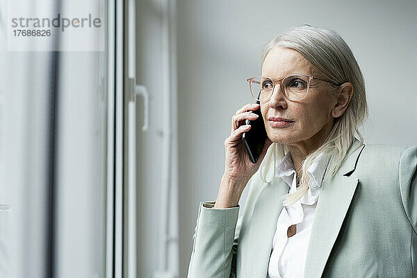 Geschäftsfrau schaut aus dem Fenster und spricht im Büro mit dem Smartphone