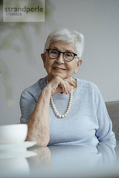 Glückliche ältere Frau mit der Hand am Kinn  die am Tisch sitzt