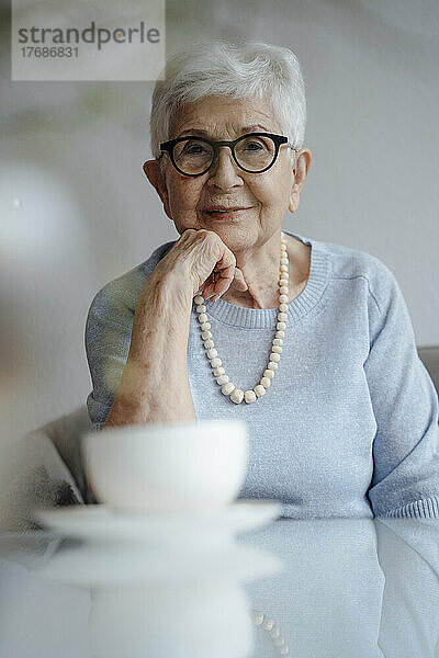 Lächelnde ältere Frau mit der Hand am Kinn  die am Tisch sitzt