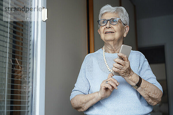Glückliche ältere Frau mit Smartphone steht am Wohnungseingang
