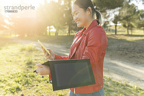 Glückliche Frau  die an einem sonnigen Tag ihr Mobiltelefon über ein Solarpanel auflädt