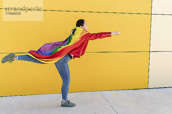 Verspielte junge Frau mit Regenbogenflaggenumhang vor gelber Wand