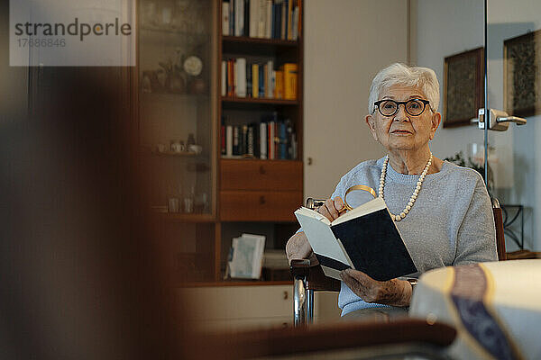 Ältere Frau mit Lupe und Buch sitzt zu Hause
