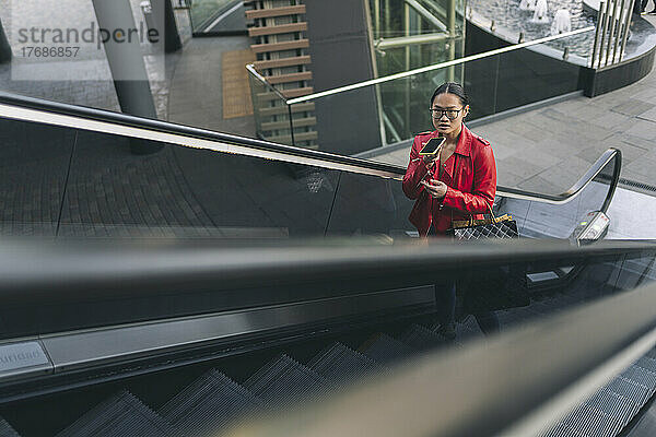 Junge Frau spricht auf der Rolltreppe über den Lautsprecher ihres Mobiltelefons