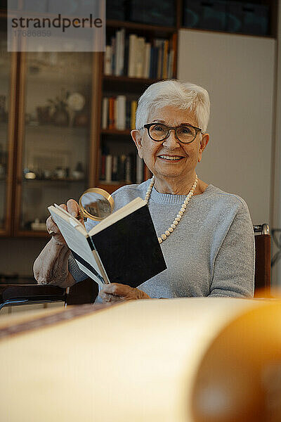 Glückliche ältere Frau mit Buch und Lupe zu Hause