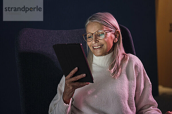 Lächelnde ältere Frau  die auf einem Stuhl sitzt und einen Tablet-PC beobachtet