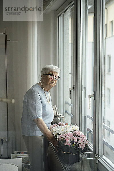 Ältere Frau steht zu Hause neben Blumen und Fenster