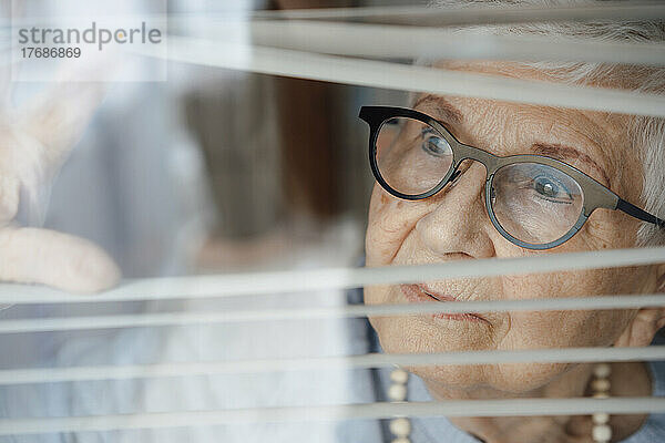 Neugierige ältere Frau schaut durch Fensterjalousien