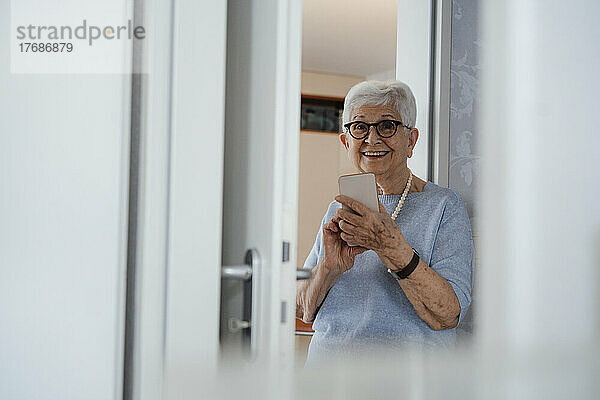 Glückliche ältere Frau mit Mobiltelefon steht zu Hause vor der Tür