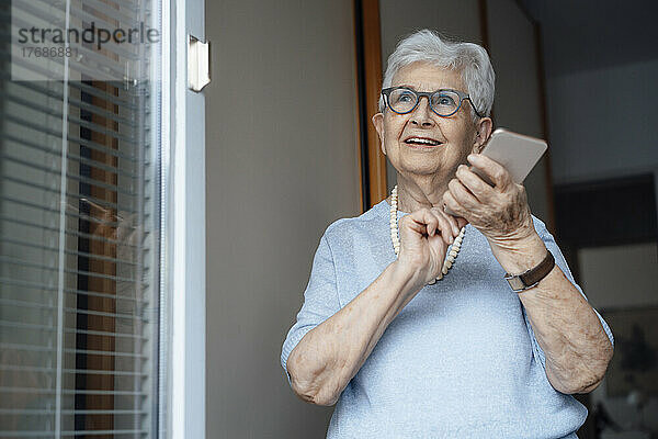 Lächelnde ältere Frau mit Smartphone steht an der Wohnungstür